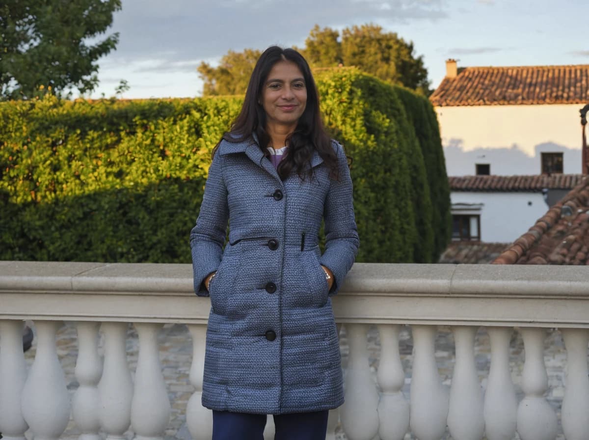 A woman standing in front of a balustrade, wearing a blue textured coat with buttons, hands in pockets. Behind them is a lush hedge and terracotta roofed houses under a soft evening sky.