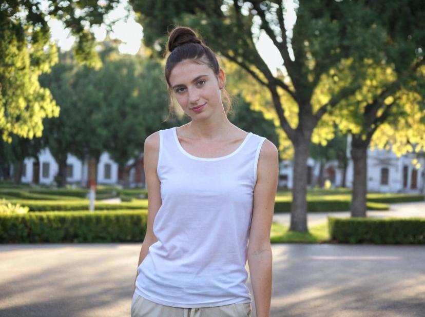 A woman with a bun hairstyle wearing a sleeveless white top and beige pants standing on a pavement with a background of a sunlit tree-lined street and green manicured lawns.