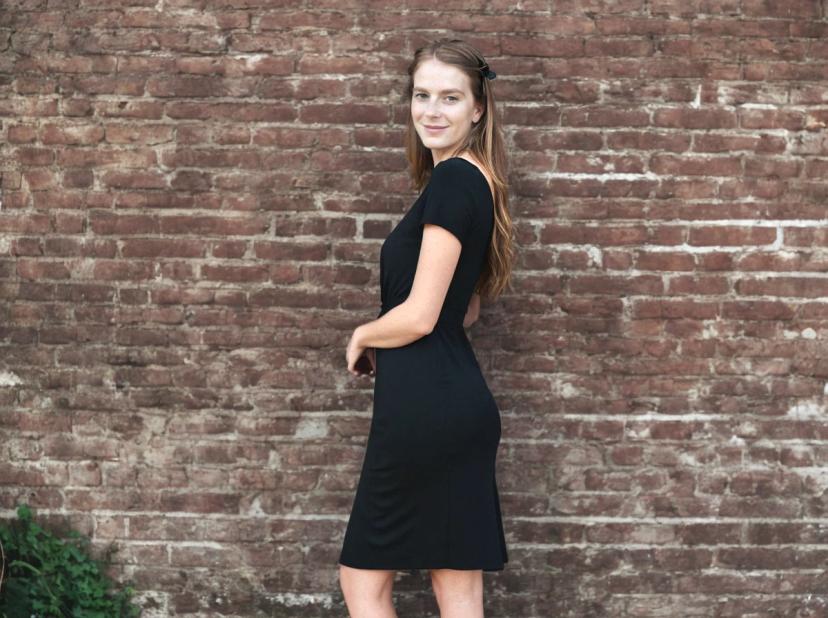 wide portrait photo of a woman with long hair tied back, wearing a short-sleeved black dress, is standing sideways in front of a textured brick wall with some foliage at the bottom.