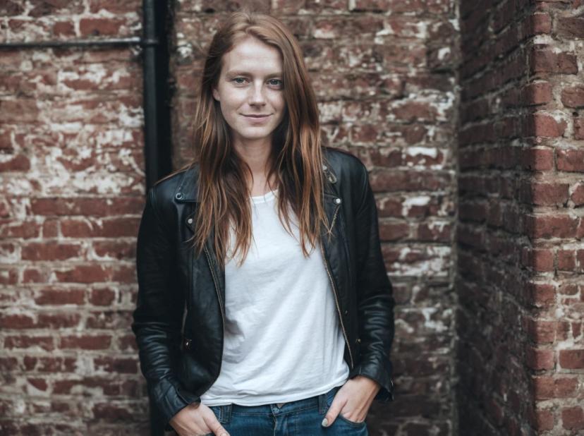 wide portrait photo of a woman wearing a black leather jacket and white t-shirt standing in front of a red brick wall.