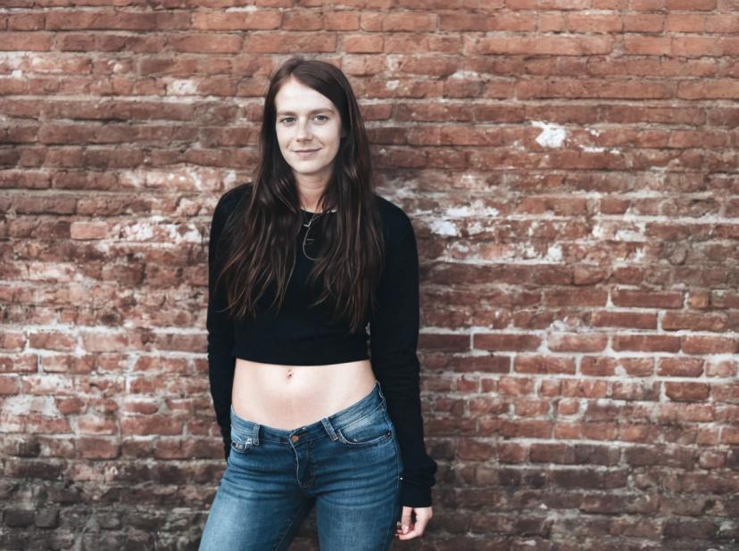 wide portrait photo of a woman wearing a black cropped top and blue jeans stands in front of a textured brick wall.