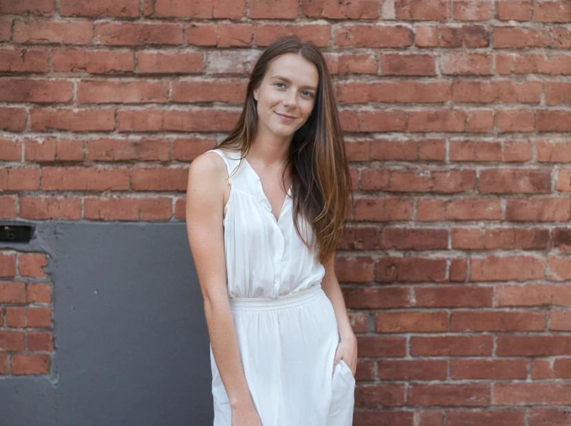 wide portrait photo of a woman wearing a white sleeveless dress standing in front of a red brick wall.