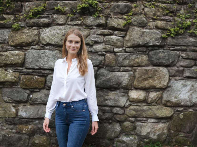 wide portrait photo of a woman standing in front of a stone wall with green plants growing between some of the stones, wearing a white blouse and blue jeans.
