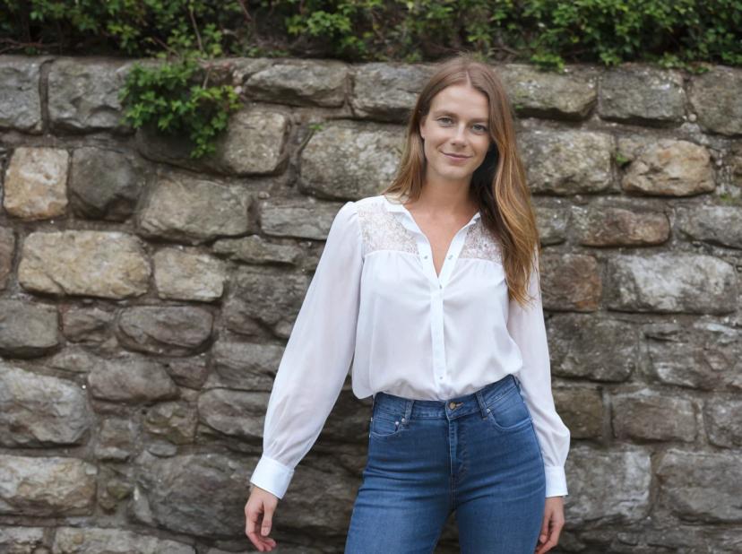 wide portrait photo of a woman wearing a white blouse with lace details and blue jeans standing in front of a stone wall with greenery.
