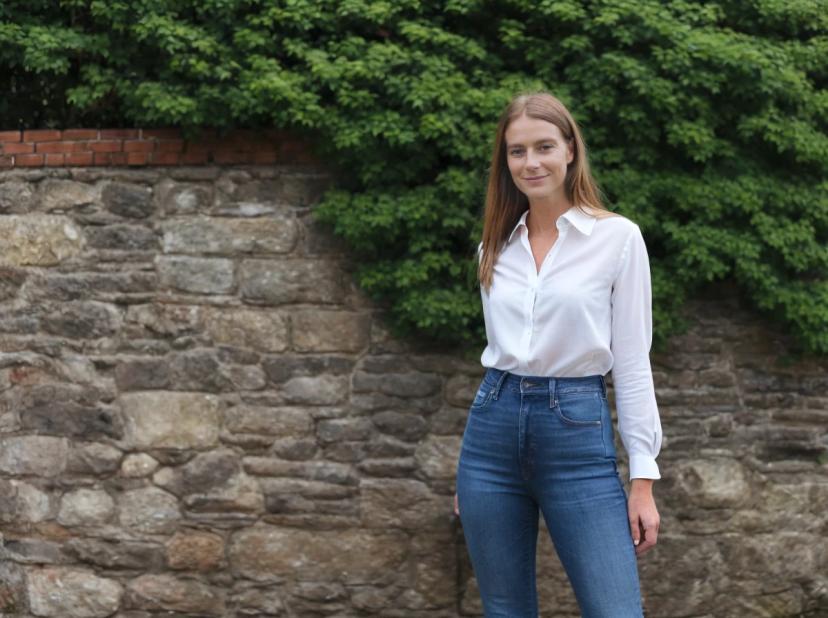 wide portrait photo of a woman wearing a white button-up shirt and blue jeans stands in front of a stone wall with dense green shrubbery on top.
