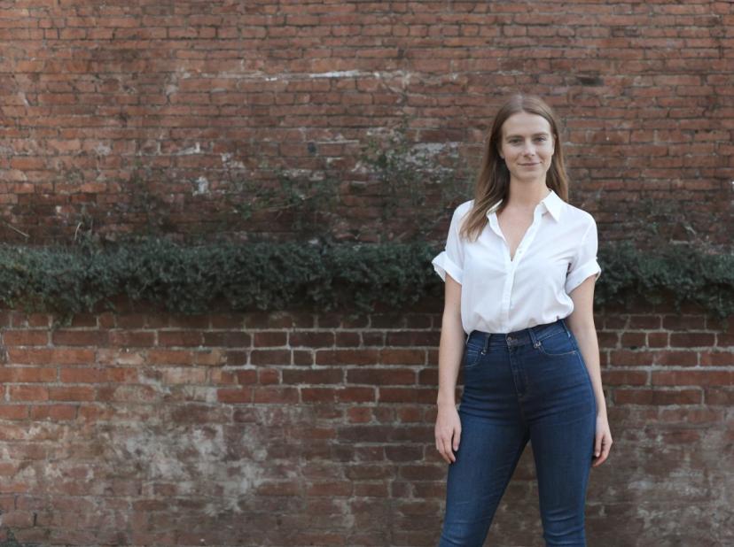 wide portrait photo of a woman wearing a white shirt tucked into blue jeans standing in front of an aged brick wall with a strip of greenery.