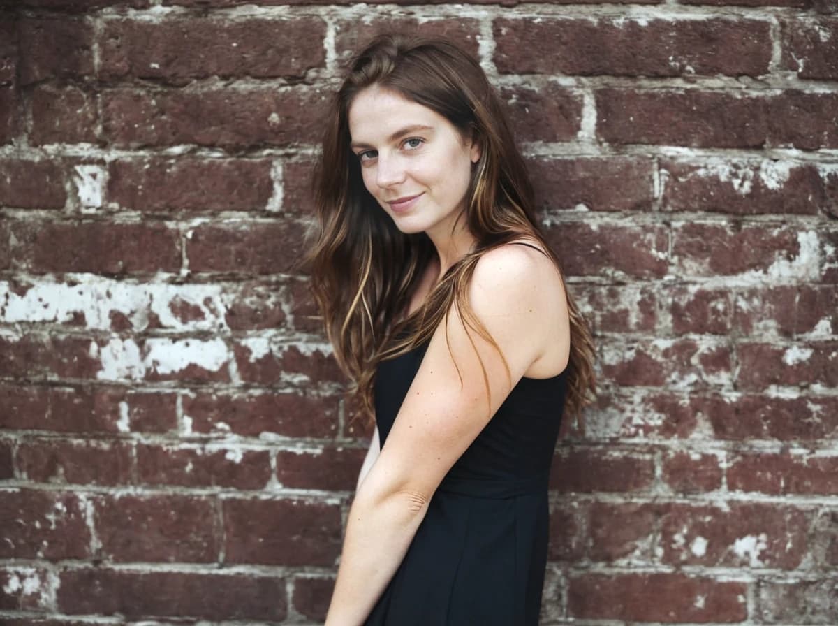 wide portrait photo of a woman with long hair wearing a black sleeveless dress stands in front of a red brick wall with white patches.