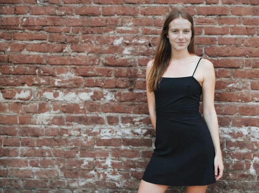 wide portrait photo of a woman in a black sleeveless dress standing in front of a textured brick wall.