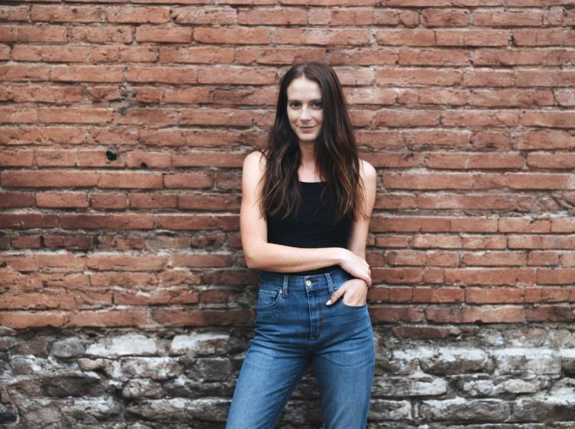 wide portrait photo of a woman with long hair wearing a black tank top and blue jeans standing in front of an aged brick wall.