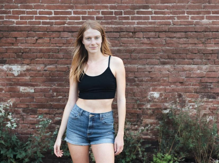 wide portrait photo of a woman standing in front of a red brick wall wearing a black tank top and blue denim shorts. There are green plants at the base of the wall.