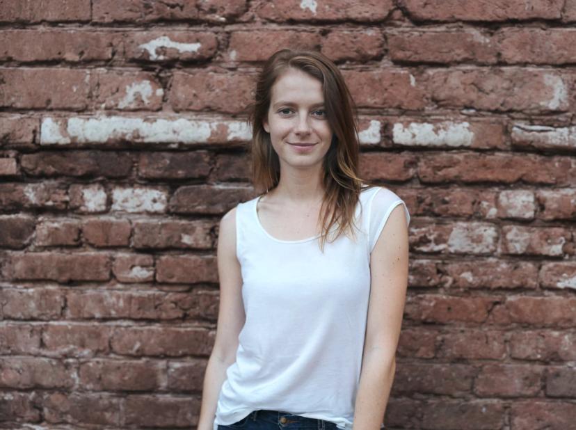 wide portrait photo of a woman standing in front of a weathered brick wall wearing a casual white sleeveless top and blue denim jeans.