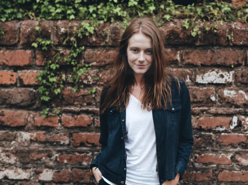 wide portrait photo of a woman wearing a dark denim jacket over a white shirt standing in front of an ivy-covered brick wall.