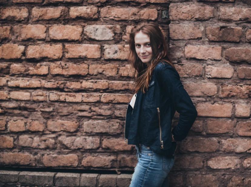 wide portrait photo of a woman in blue denim jeans and a black jacket stands in profile against a rustic brick wall.