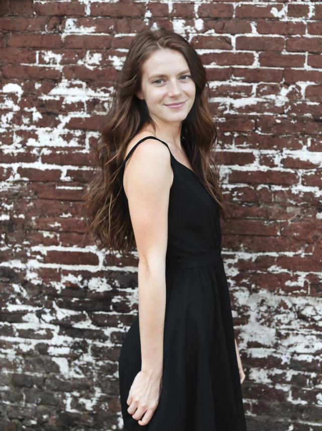 portrait photo of a woman with long hair standing in front of a weathered brick wall wearing a black sleeveless dress.