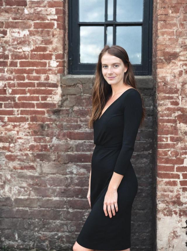 portrait photo of a woman wearing a black dress stands in front of an old brick wall with a window featuring black frames.