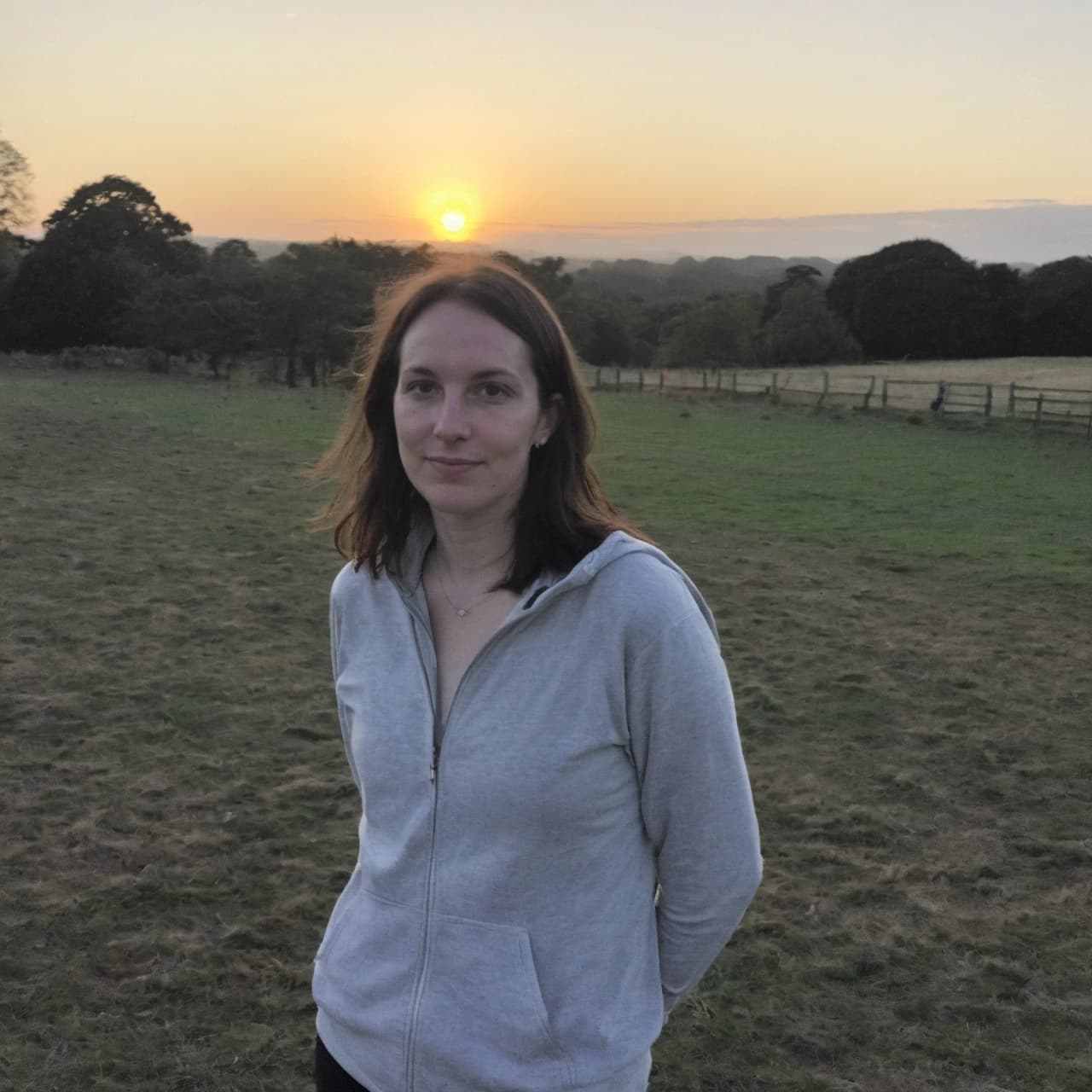 A woman in a grey zip-up hoodie stands in a grassy field at sunset, with trees and a fence in the background.