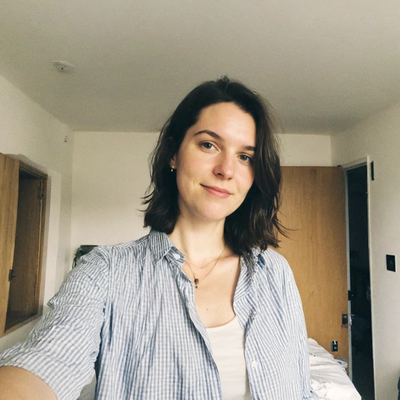 A young woman with shoulder-length brown hair and a subtle smile wears a blue and white striped button-up shirt over a white top, standing in a room with light-colored walls, a partially visible wooden door, and a white ceiling.