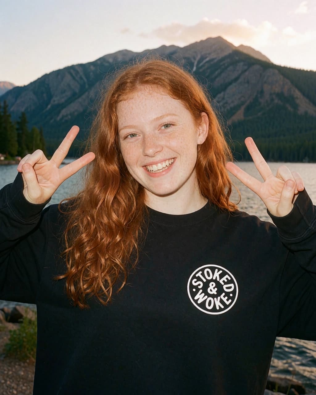 A young woman with long red hair and freckles smiles while making peace signs with both hands in front of a lake and mountains. They are wearing a black long-sleeved shirt with a circular white logo that reads STOKED & WOKE on the chest. The setting is outdoors during daytime with natural lighting.