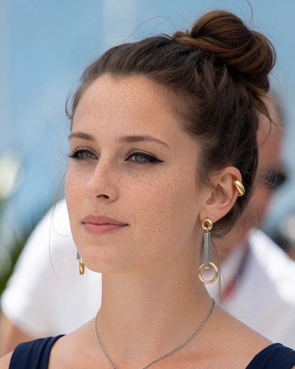 A woman with fair skin and visible freckles on her nose and cheeks is looking toward the left side of the frame. Her brown hair is styled in a high bun. She is wearing winged eyeliner, a thin silver necklace, and gold-toned earrings featuring a small hoop, a rectangular bar, and a larger hoop. She also wears a gold ear cuff on her left ear and is dressed in a dark blue top. The background is slightly out of focus, showing a blurred outdoor setting.