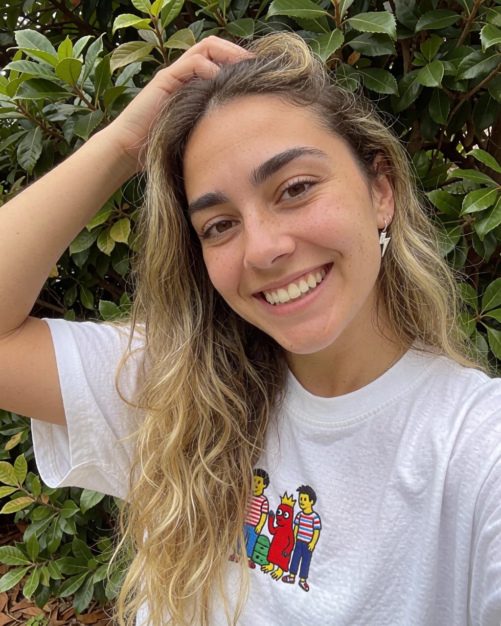 A young woman with blonde-highlighted brown hair is smiling at the camera while holding one hand to the back of her head. She has freckles on her face and is wearing a white t-shirt with a small graphic print of three cartoon figures on the chest. She is also wearing a small silver lightning bolt earring in her left ear. Behind her is a dense, leafy green shrub.
