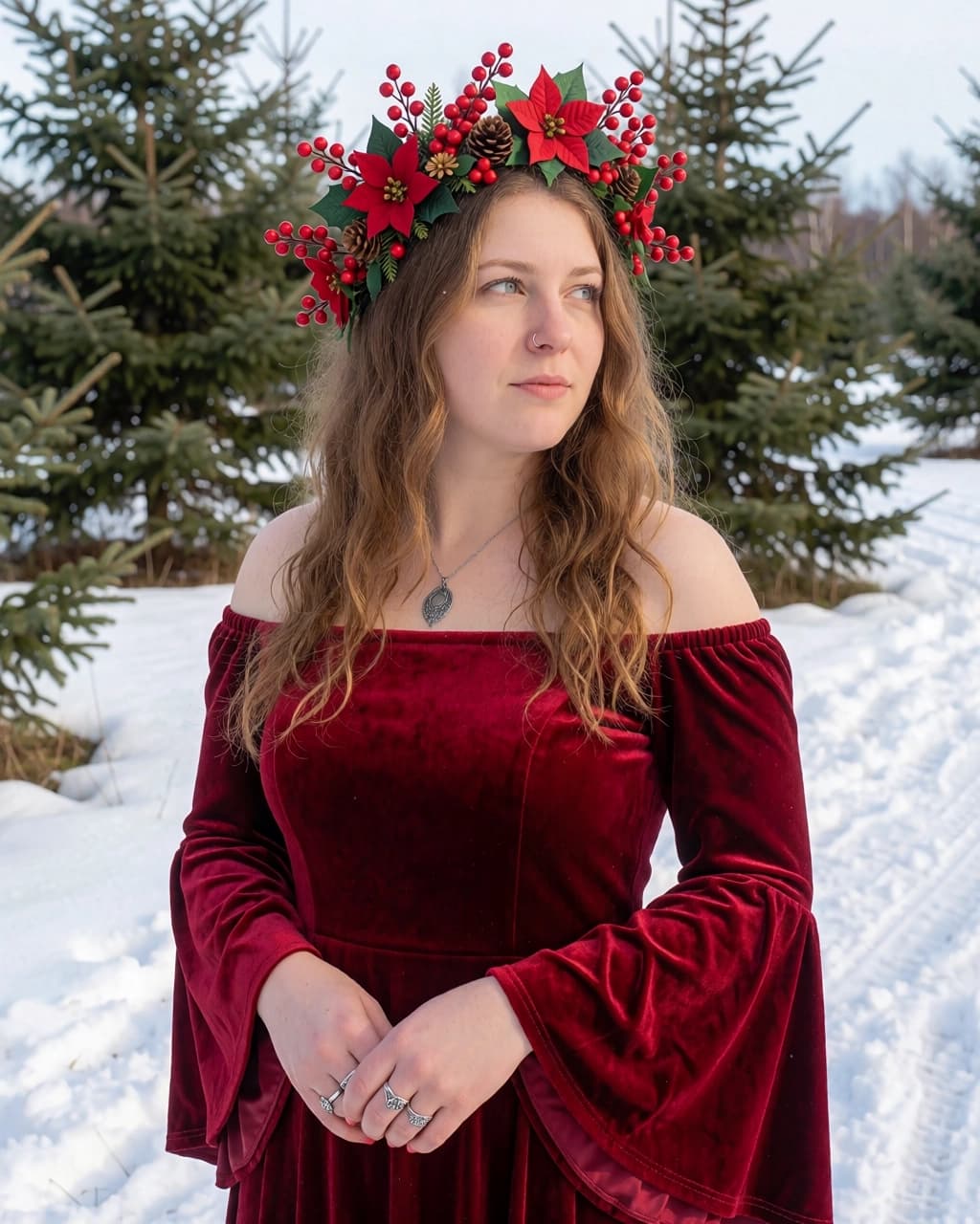 A young woman with wavy brown hair is wearing a deep red velvet off-the-shoulder dress with bell sleeves. She has a flower crown on her head featuring red poinsettias, red berries, and small pinecones. She is wearing a silver pendant necklace and rings on her fingers. She is standing outdoors in a snow-covered area with evergreen trees in the background.