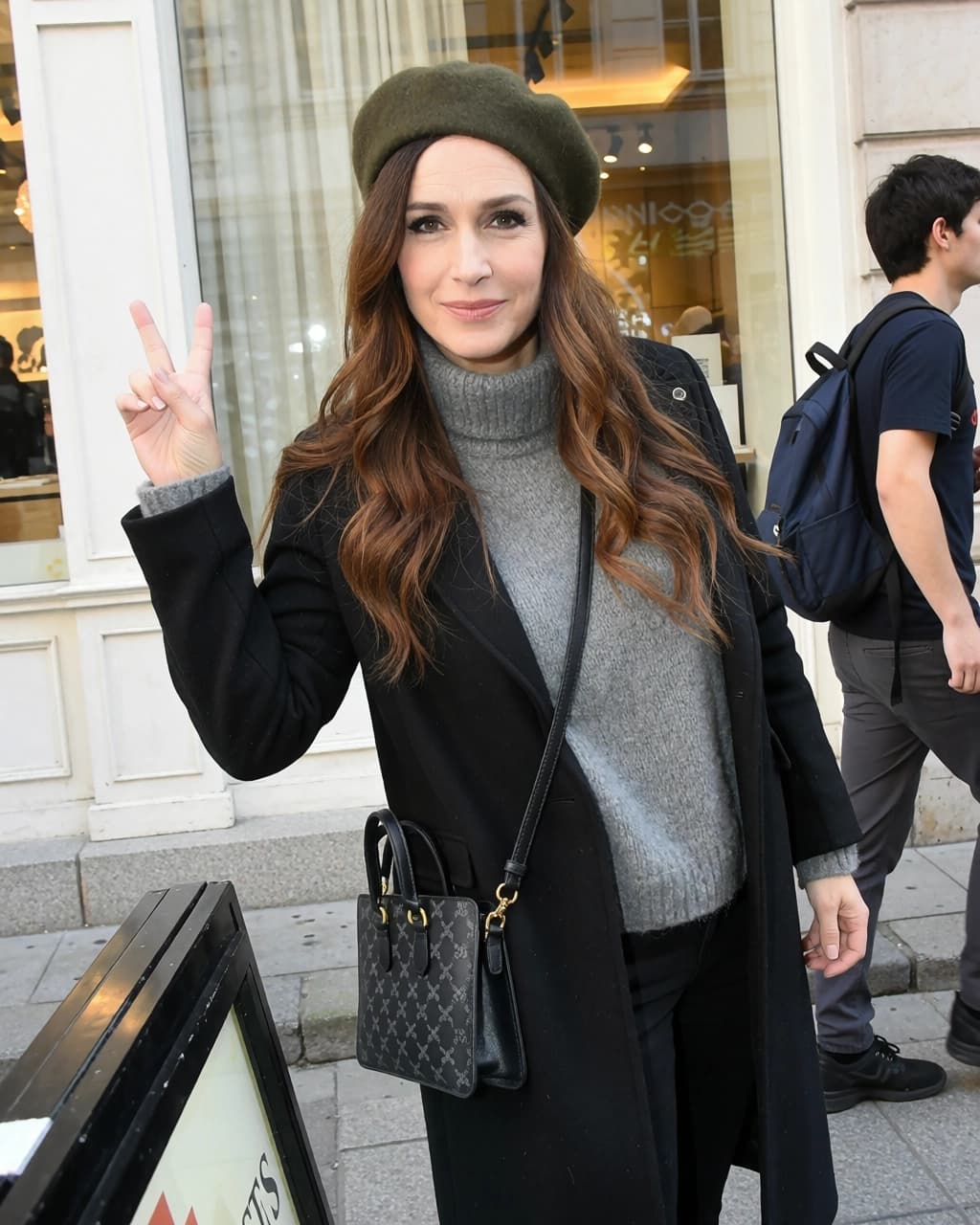 A woman with long brown hair wears an olive green beret, a grey turtleneck sweater, and a black coat while making a peace sign with her right hand. She carries a small black patterned handbag with a shoulder strap across her body. She stands in front of a storefront window with a white frame. To her right, a man wearing a dark t-shirt and carrying a blue backpack walks past her.
