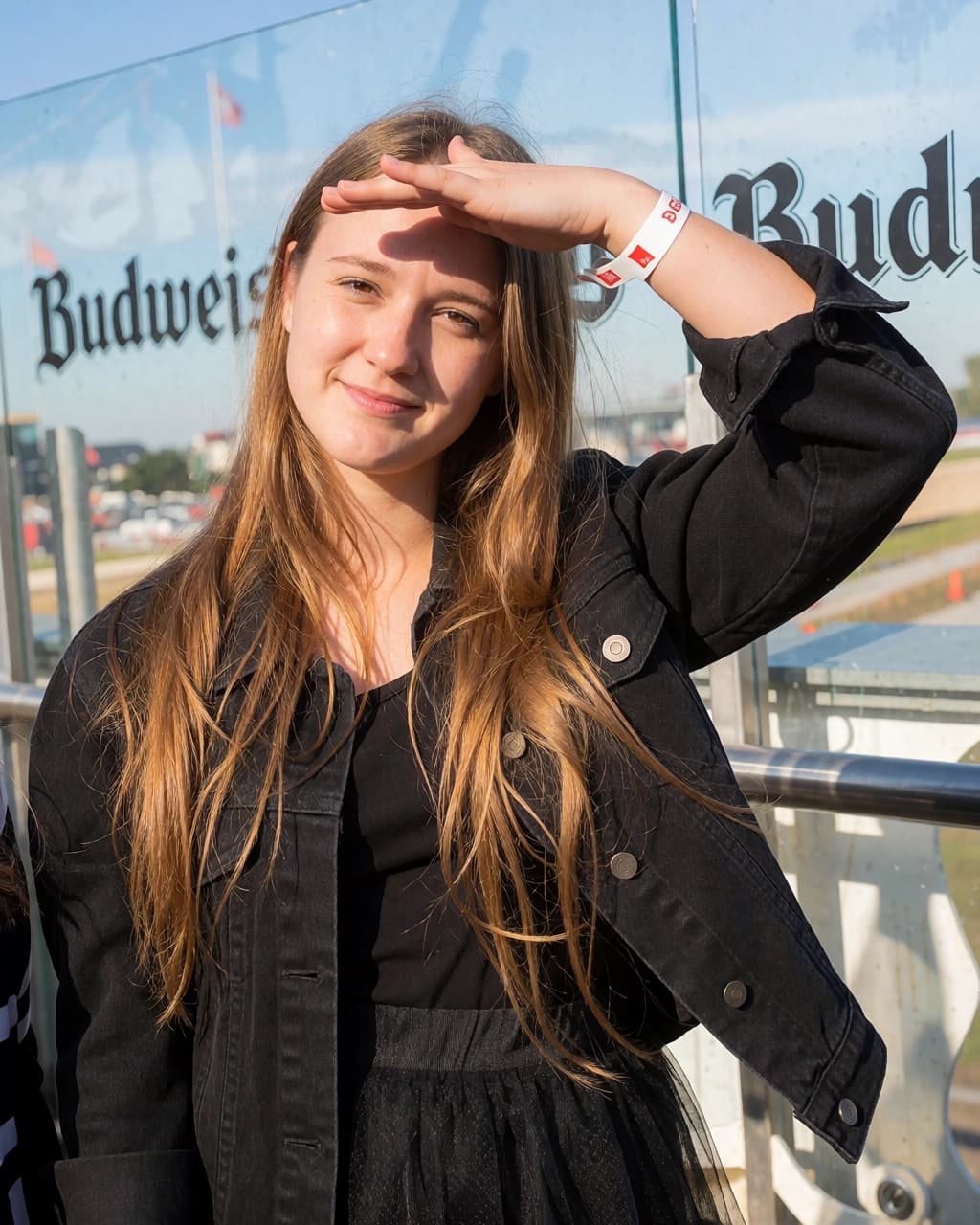 A young woman with long light brown hair is standing outdoors in front of a glass wall with the word Budweiser printed on it. She is wearing a black denim jacket over a black shirt and skirt. She holds her left hand up to her forehead as a visor to block the sun, revealing a white wristband on her arm. She is looking directly at the camera with a subtle smile.