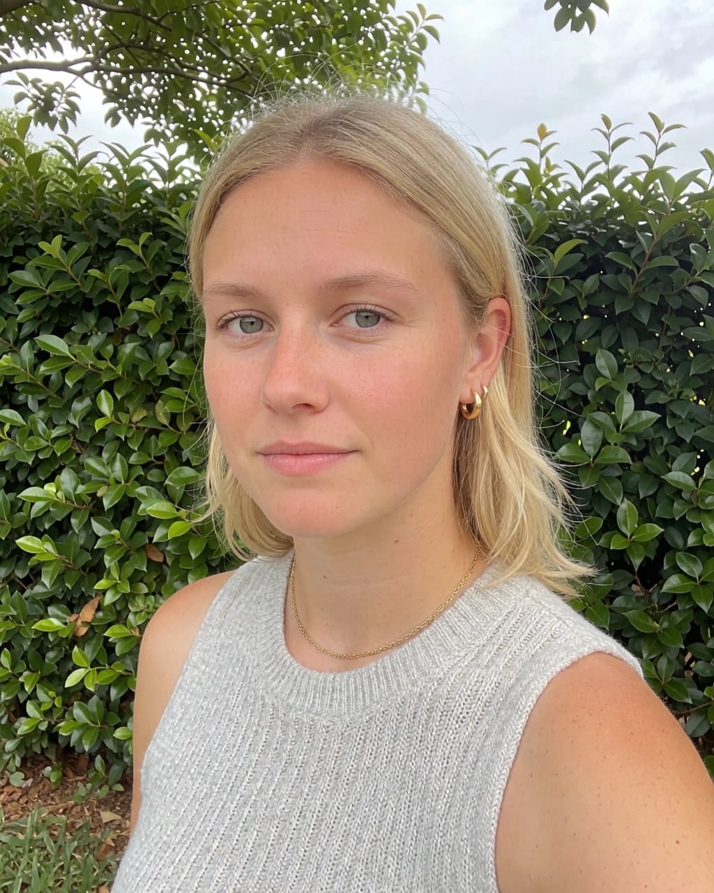 A blonde woman with her hair parted in the middle looks directly at the camera. She is wearing a light gray sleeveless ribbed top, a thin gold chain necklace, and small gold hoop earrings in her left ear. A dense green hedge serves as the background.