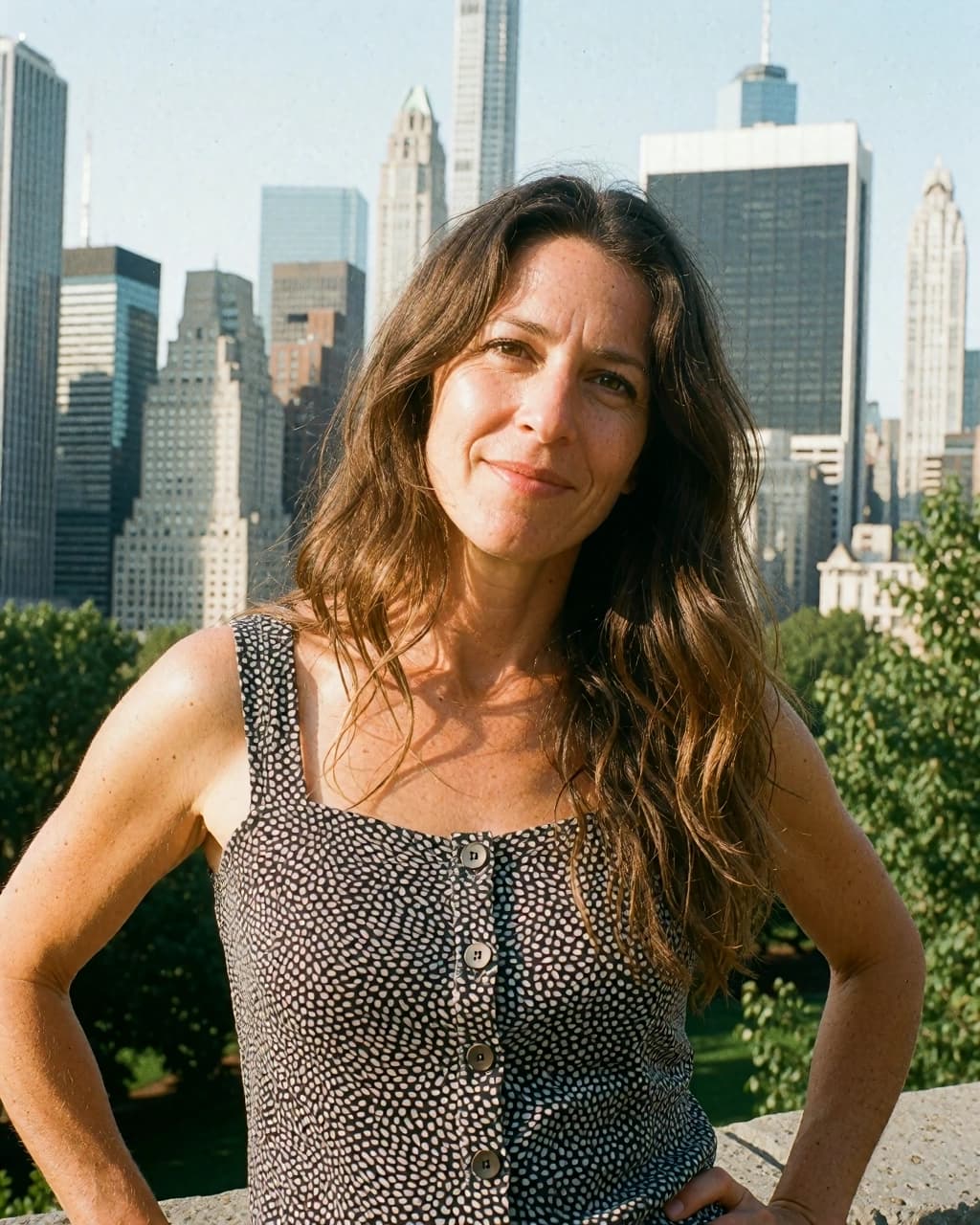 A woman with long, brown wavy hair stands in front of a city skyline with buildings and green trees. She is wearing a black sleeveless top with white polka dots and four buttons down the front. Her hands are placed on her hips and she is looking directly at the camera with a slight smile. The background is a dense urban scene with multiple skyscrapers visible under a clear, bright sky.