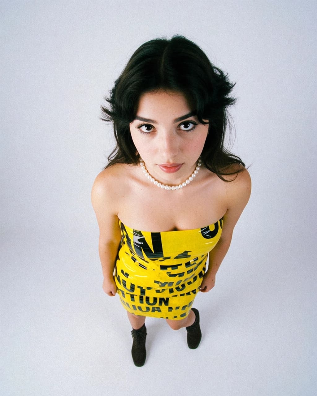 A woman with dark hair stands against a plain white background, viewed from a high angle. She wears a strapless dress made of yellow caution tape with black text, a white pearl necklace, and dark ankle boots. She looks directly up at the camera with a neutral expression.