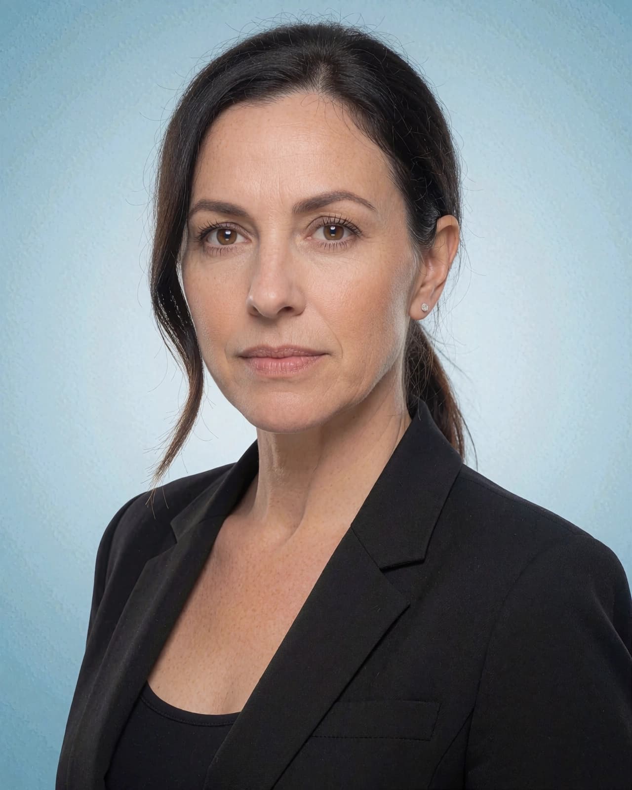 A medium shot of a woman with brown hair pulled back into a ponytail, facing forward against a light blue background. She is wearing a black blazer over a black top and a small earring in her left ear. Her hair is parted in the center, and a few strands frame her face. She has brown eyes and is looking directly at the camera with a neutral expression.