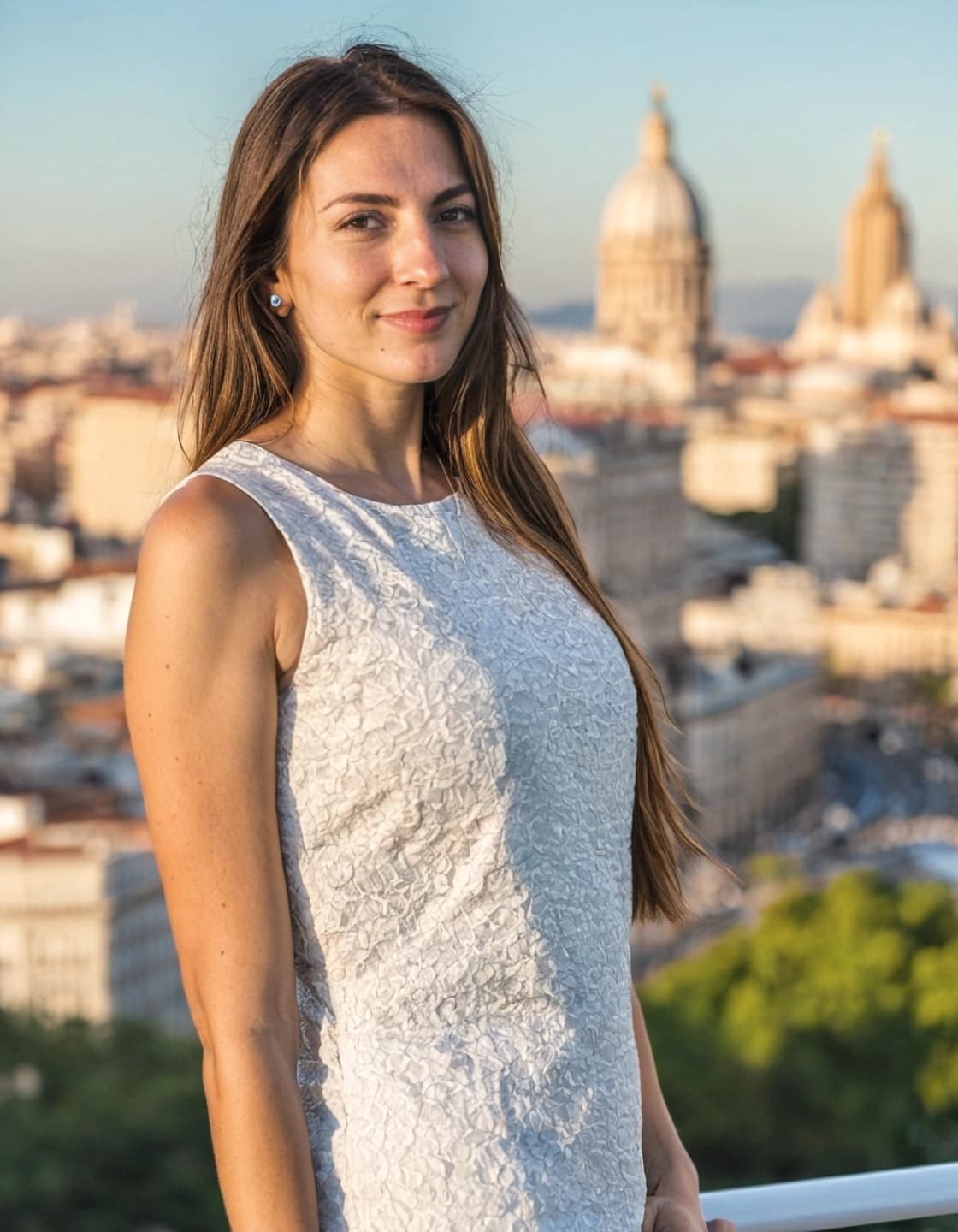 A young woman with long, dark brown hair and a slight smile stands outdoors with a city panorama in the blurred background. She is wearing a white sleeveless top with a textured pattern and small stud earrings. In the background, domed buildings and other structures are visible under a clear sky.